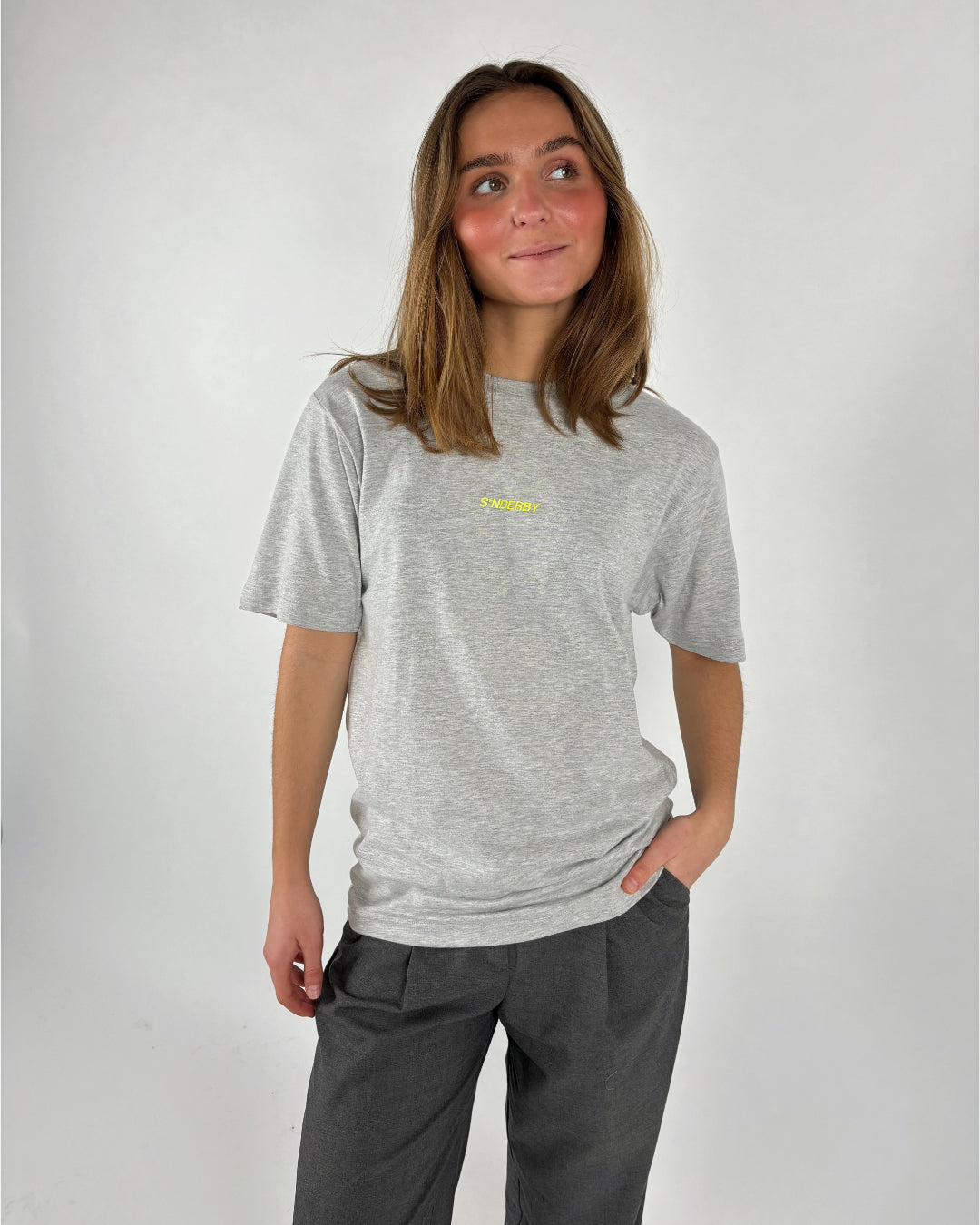 En person iført en grå T-shirt med teksten "S'NOEBY" står afslappet med hånden i lommen på et par mørke bukser, mod en lys baggrund.