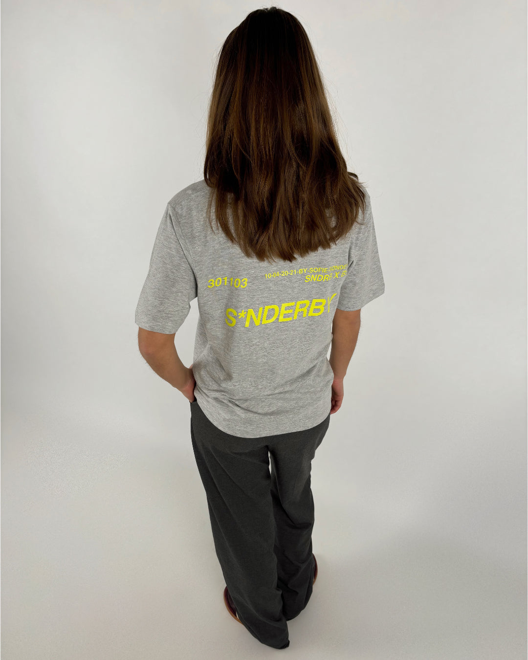 En person med langt hår står med ryggen til iført grå T-shirt og mørke bukser. Tekst på T-shirten: "301103 10-04-2021 BY SOFIE SØNDERBY SNDRBX STUDIO SØNDERBY." Baggrund er enkel og lys.