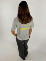 En person med langt hår står med ryggen til iført grå T-shirt og mørke bukser. Tekst på T-shirten: "301103 10-04-2021 BY SOFIE SØNDERBY SNDRBX STUDIO SØNDERBY." Baggrund er enkel og lys.