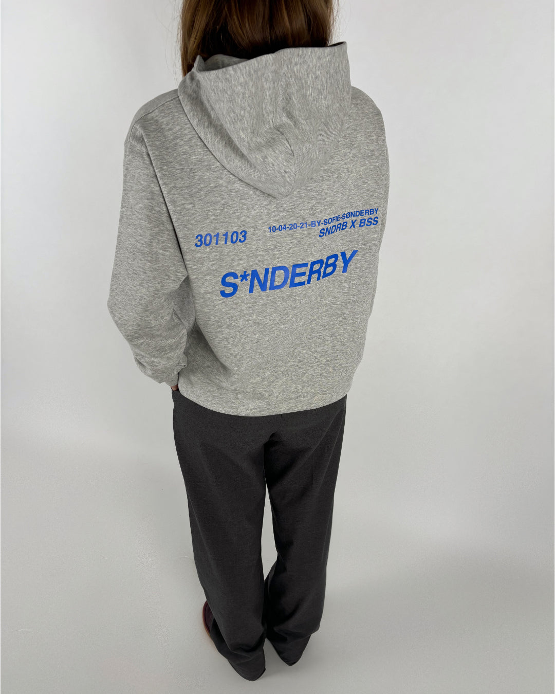 En person iført en grå hættetrøje står med ryggen til. På ryggen står teksten: "301103", "10-04-20-21-BY-SOFIE SØNDERBY", "SNDRB X BSS", "S*NDERBY". Baggrunden er hvid.