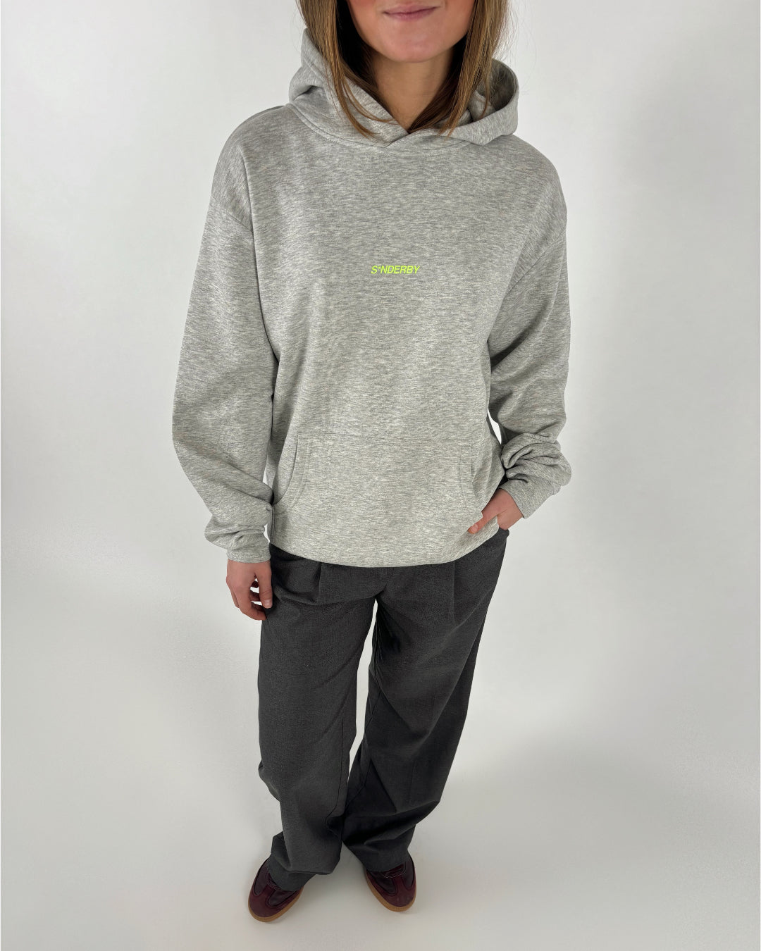 Kvinde i en grå hoodie med teksten "SØNDERBY" står med hænderne i lommen, iført grå bukser og brune sko, på en neutral baggrund.