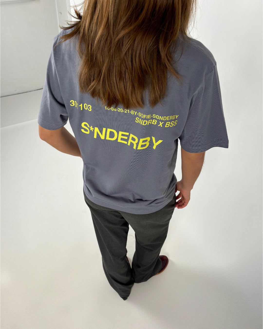 En person med langt hår står i et hvidt rum, iført en grå T-shirt med gul tekst: "30 / 103, 16-04-20-21-BY-SOFIE-SØNDERBY, SNDRB X BSS, S*NDERBY".