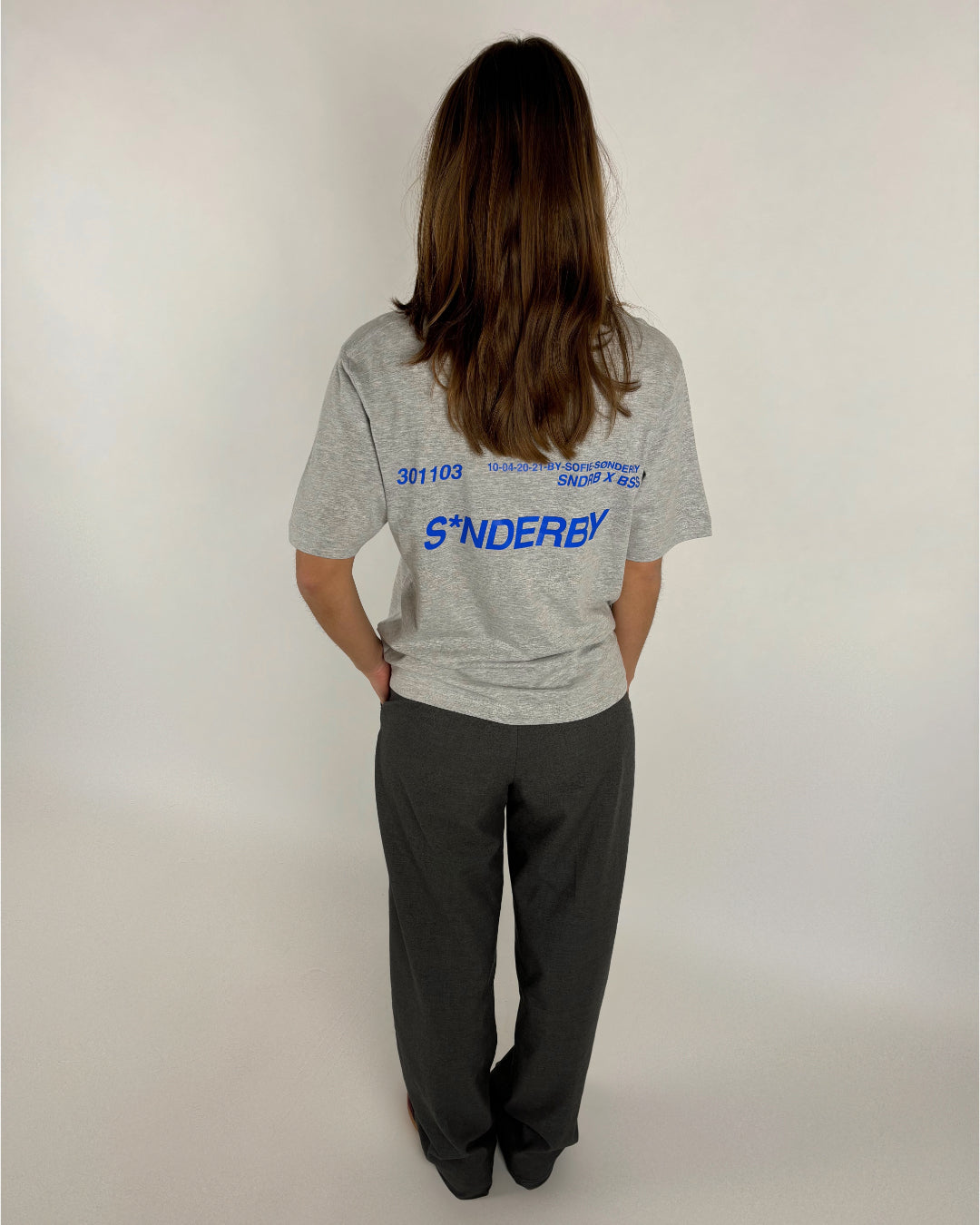 En person med langt hår bærer en grå T-shirt med blå tekst "SØNDERBY 301103" og yderligere tekst i et studie med hvid baggrund.