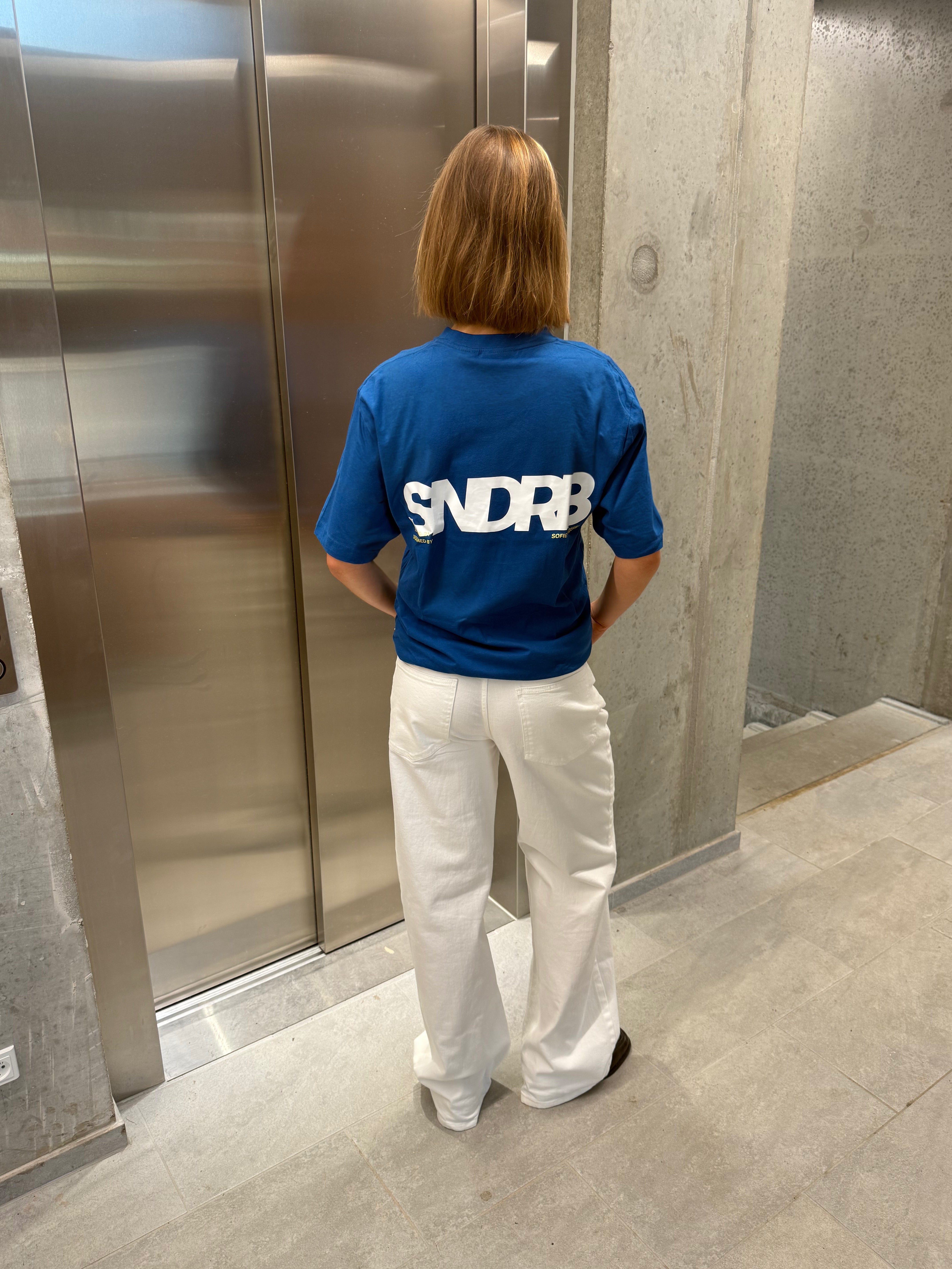 Stemningsbillede af pige der bærer en kongeblå t-shirt med hvidt SNDRB tryk og hvide NANNA jeans.  Fra BySofieSønderby