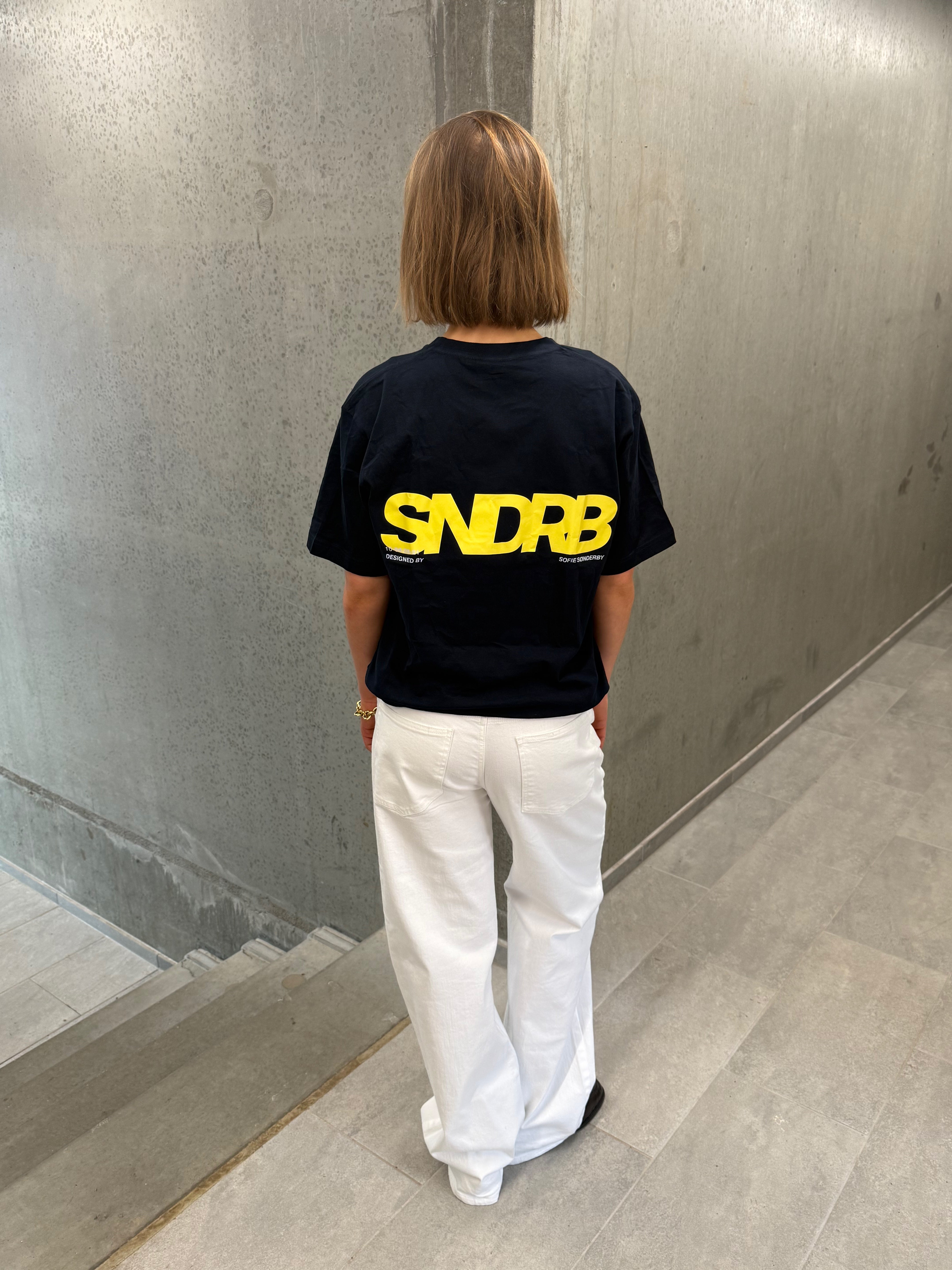 Stemningsbillede af pige der bærer en navy t-shirt med gul SNDRB tryk og hvide NANNA jeans.  Fra BySofieSønderby