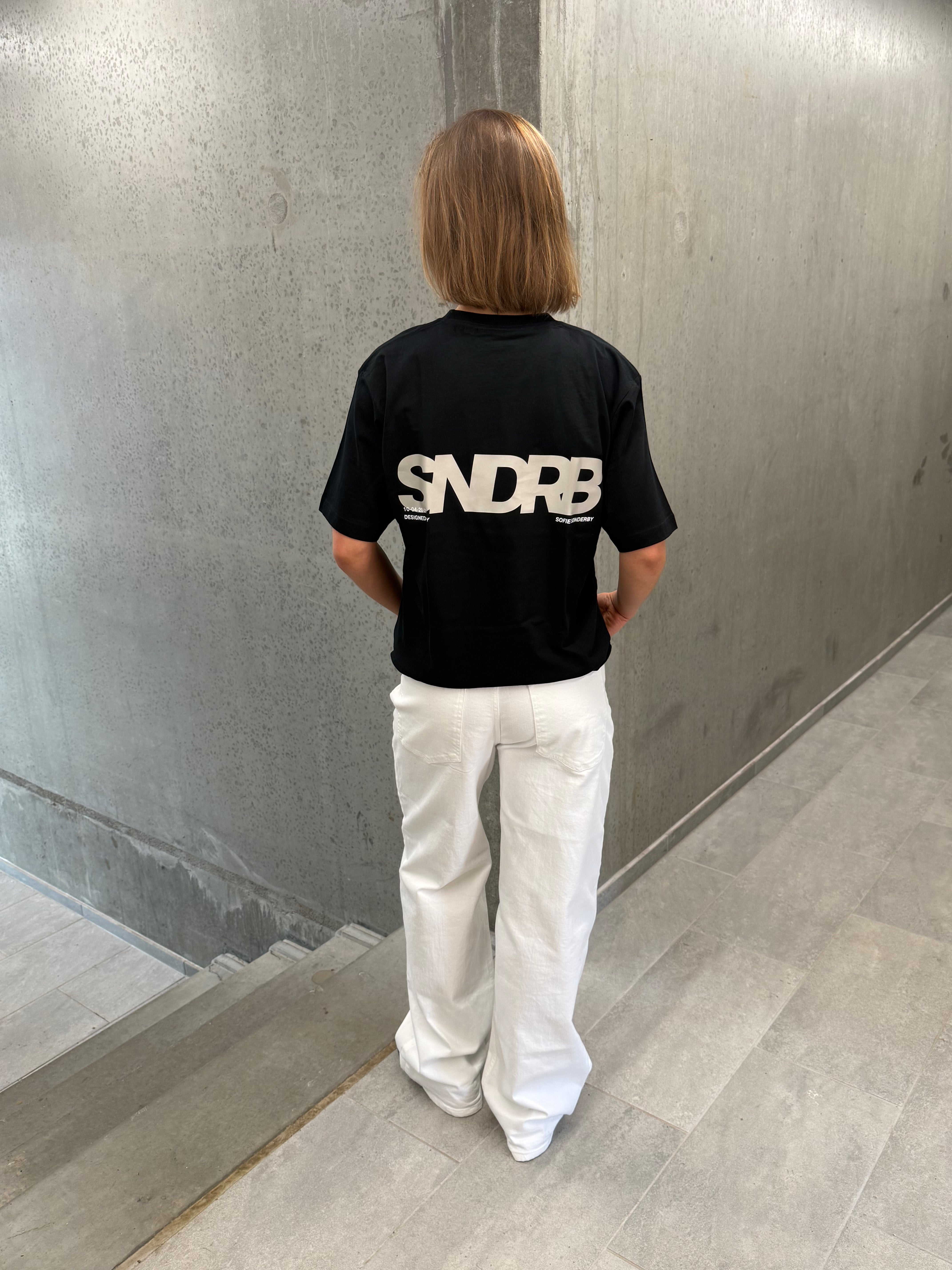 Stemningsbillede af pige der bærer en sort t-shirt med beige SNDRB tryk og hvide NANNA jeans.  Fra BySofieSønderby