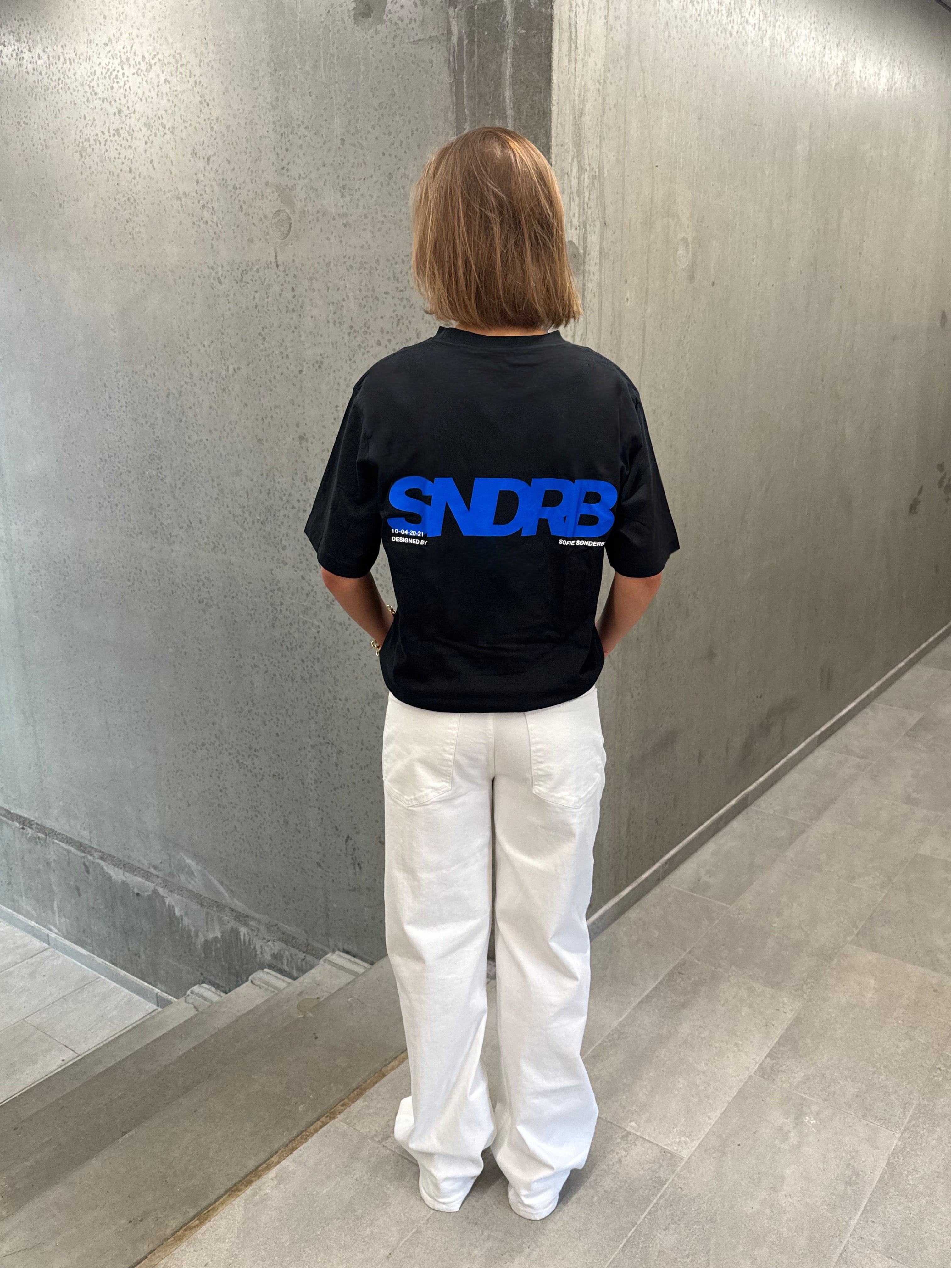 Stemningsbillede af pige der bærer en sort t-shirt med blå SNDRB tryk og hvide NANNA jeans.  Fra BySofieSønderby