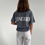 Modelfoto af pige som bærer en SNDRB tee i farven stone, samt et par hvide jeans. Alt fra By Sofie Sønderby