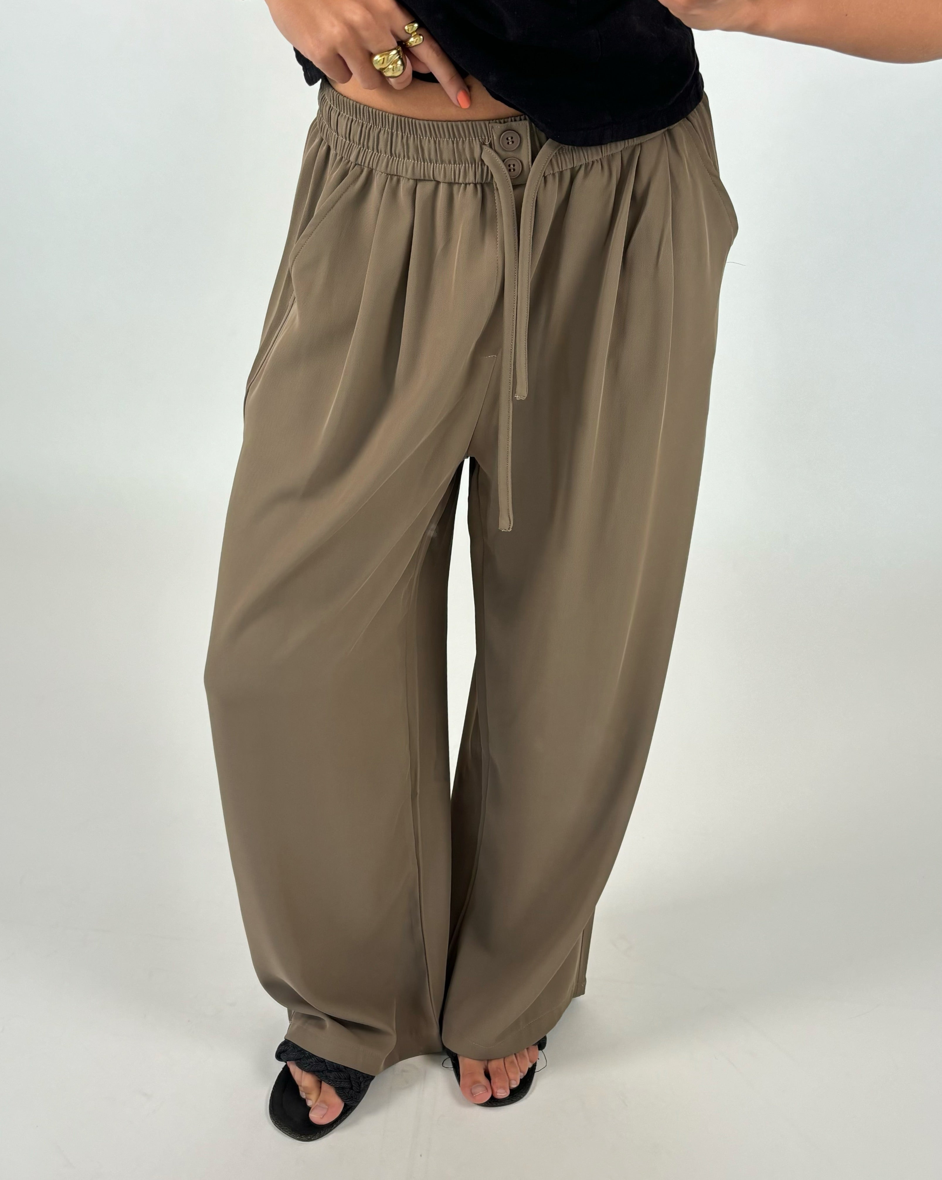 Lys brune bukser med elastisk talje og snor ses på en person med sort top og sorte sandaler, der står mod en neutral baggrund. Personen har guldringe på fingrene.