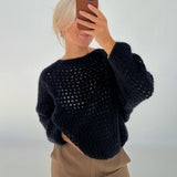 En person iført en sort, hullet sweater tager en selfie med en mobiltelefon i en lys baggrund. Personen har lyst hår og afslappede brune bukser.