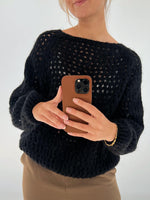 Person tager et spejlbillede med en smartphone. Personen bærer en sort strikket sweater og en brun bukser, med et enkelt guldsmykke om håndleddet. Baggrunden er enkel og lys.