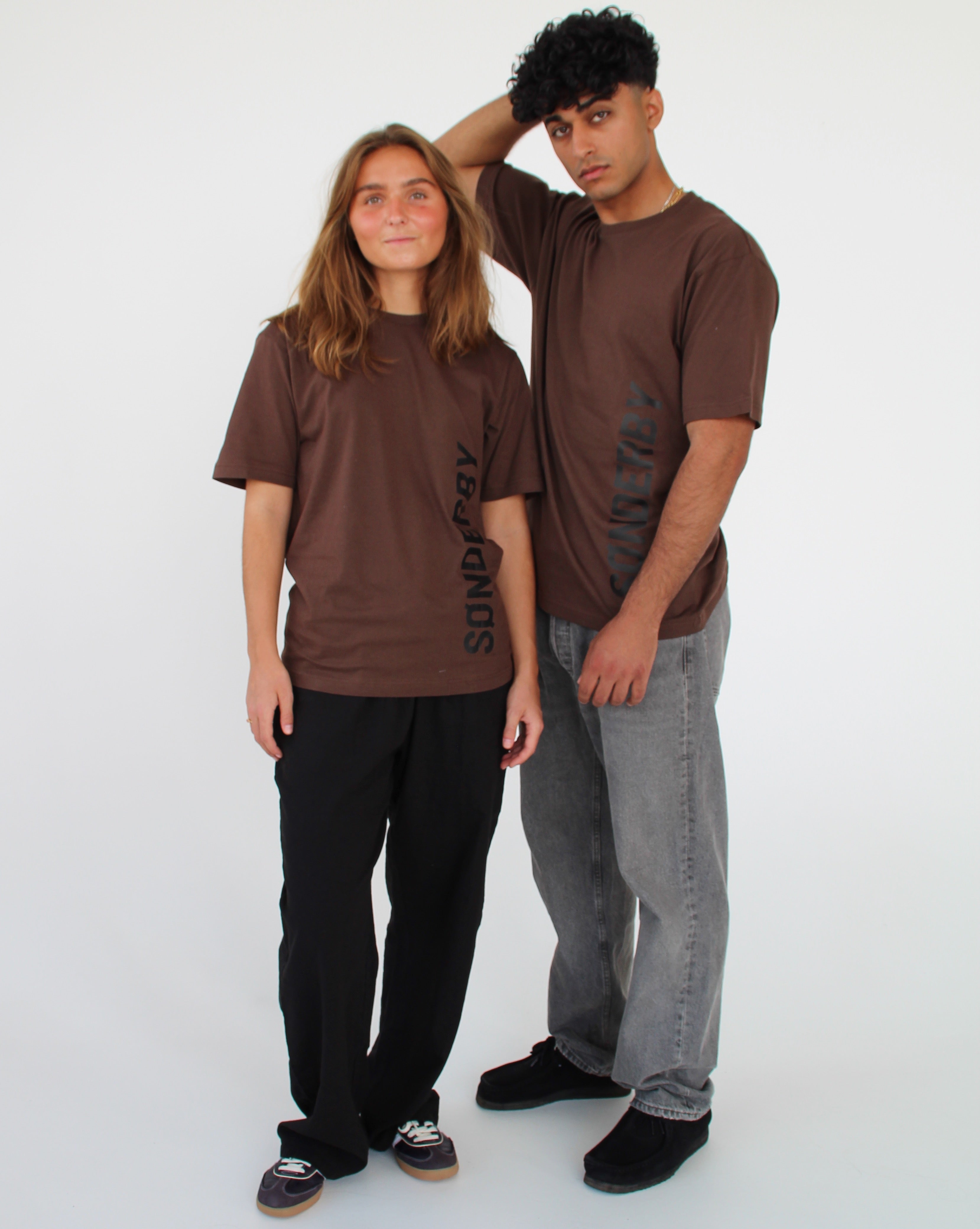 To personer står sammen iført brune T-shirts med teksten "SØNDERBY." De står i en neutral, hvid baggrund, med afslappede jeans og bukser.