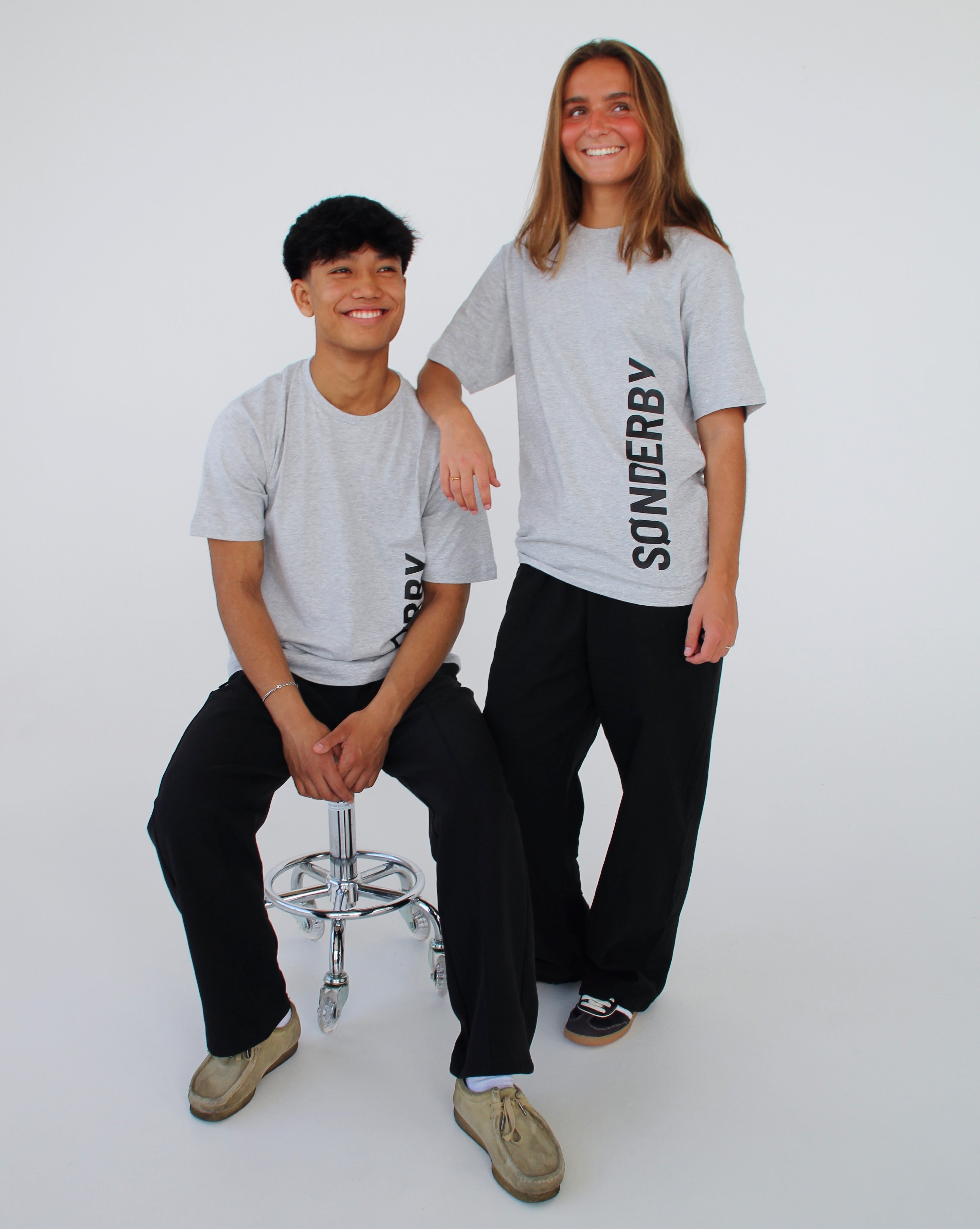 To personer i grå T-shirts med teksten "SØNDERBY". Den ene sidder på en metalstol, den anden står ved siden af. De smiler i et hvidt studie.