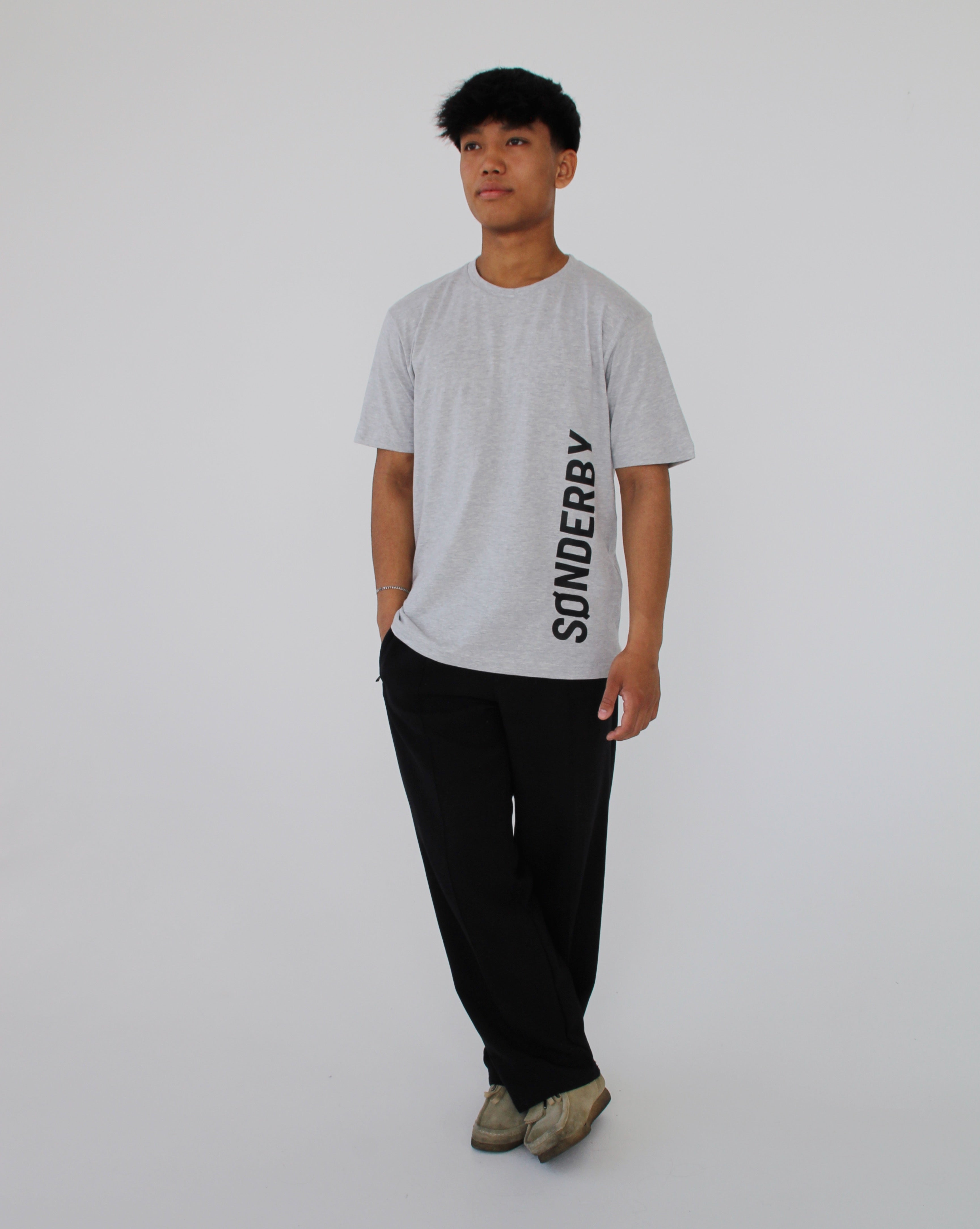 Person står iført en grå t-shirt med teksten "SØNDERBY", sorte bukser og beige sko mod en neutral baggrund.