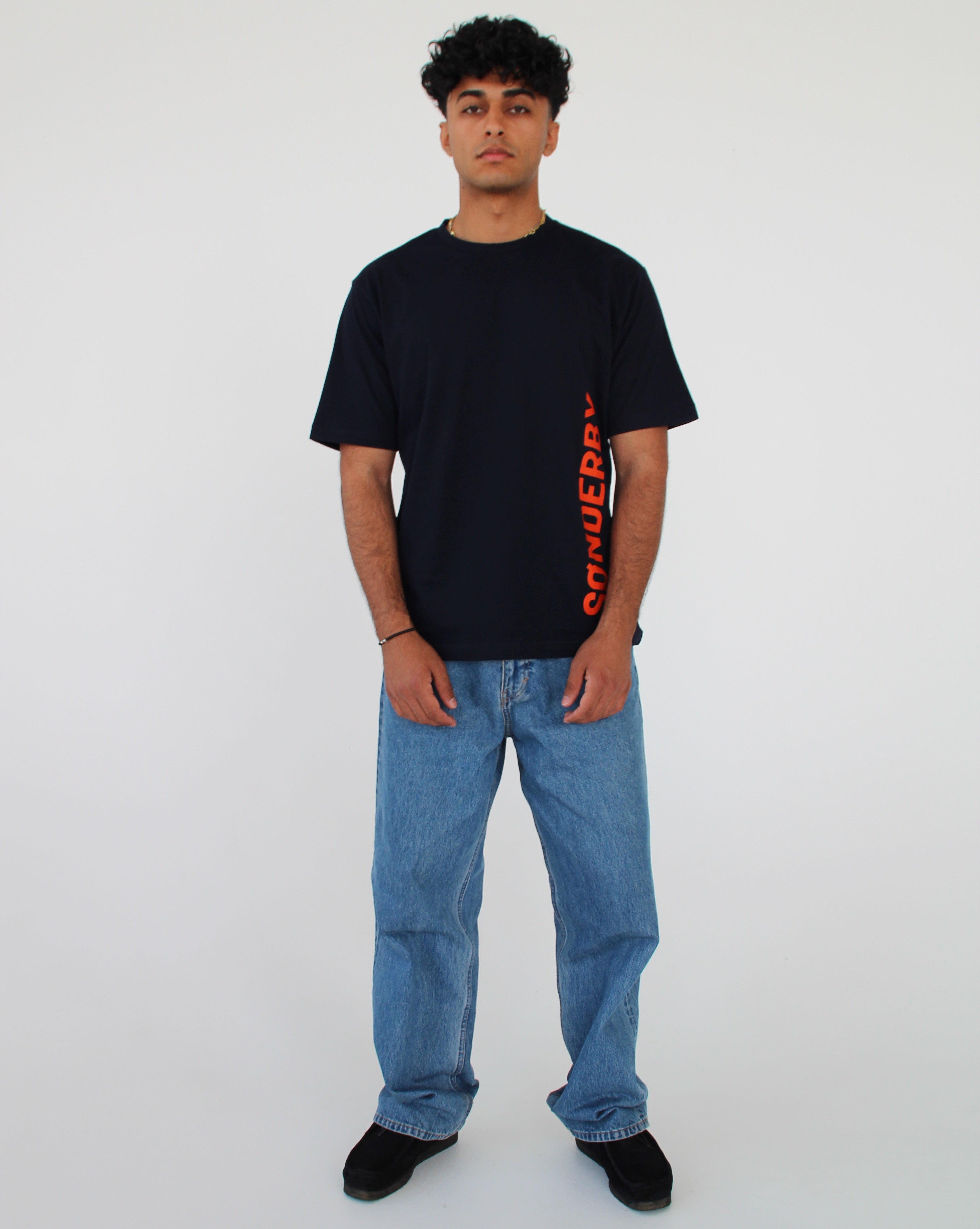 En person står i et hvidt rum, iført en sort T-shirt med orange tekst "CONQUER" og lyseblå jeans. Personen ser fremad med armene afslappet ned langs siderne.