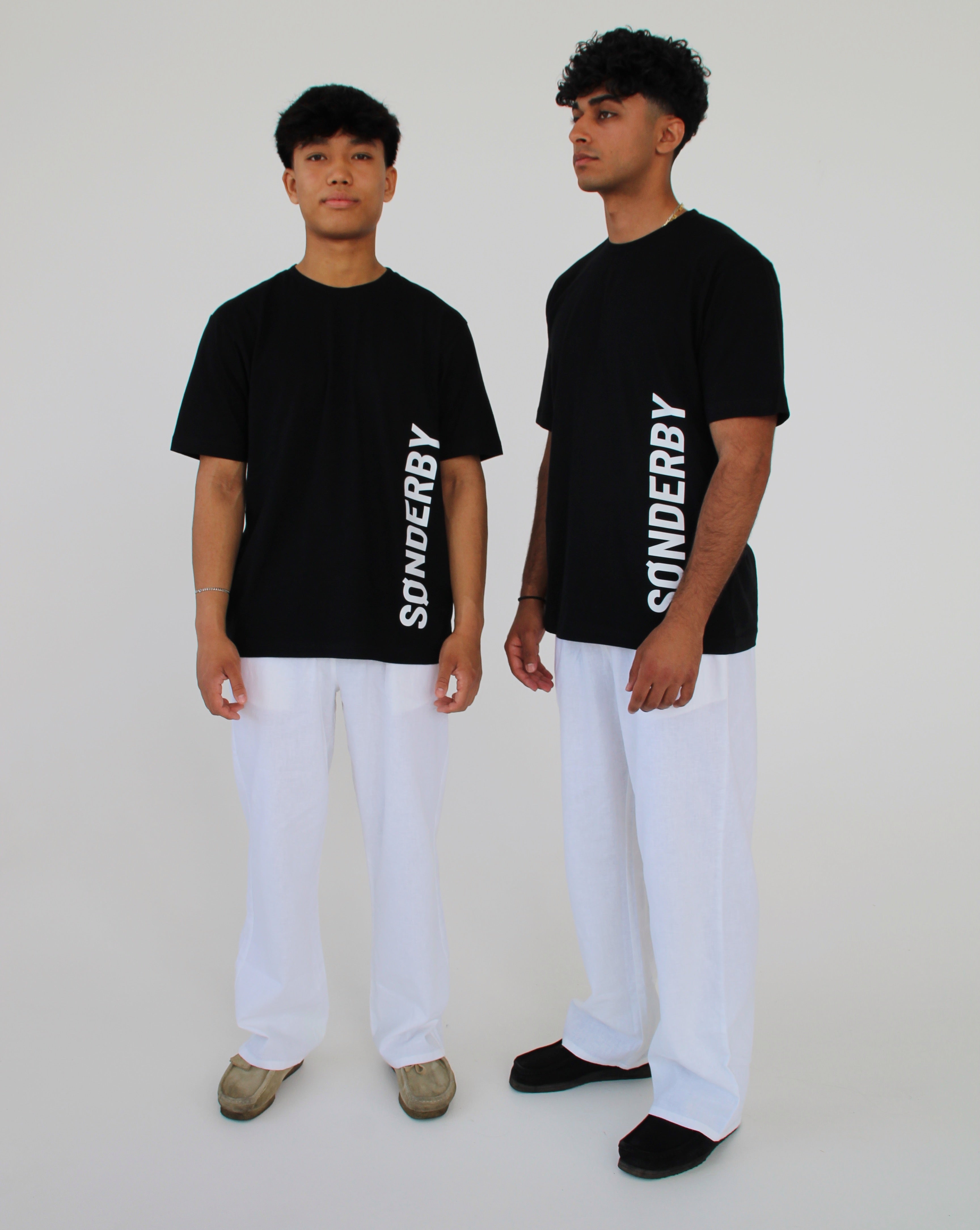 To personer iført sorte T-shirts med teksten "SØNDERBY" står i et lyst rum, og har hvide bukser på. De ser fremad, i et enkelt og minimalistisk miljø.