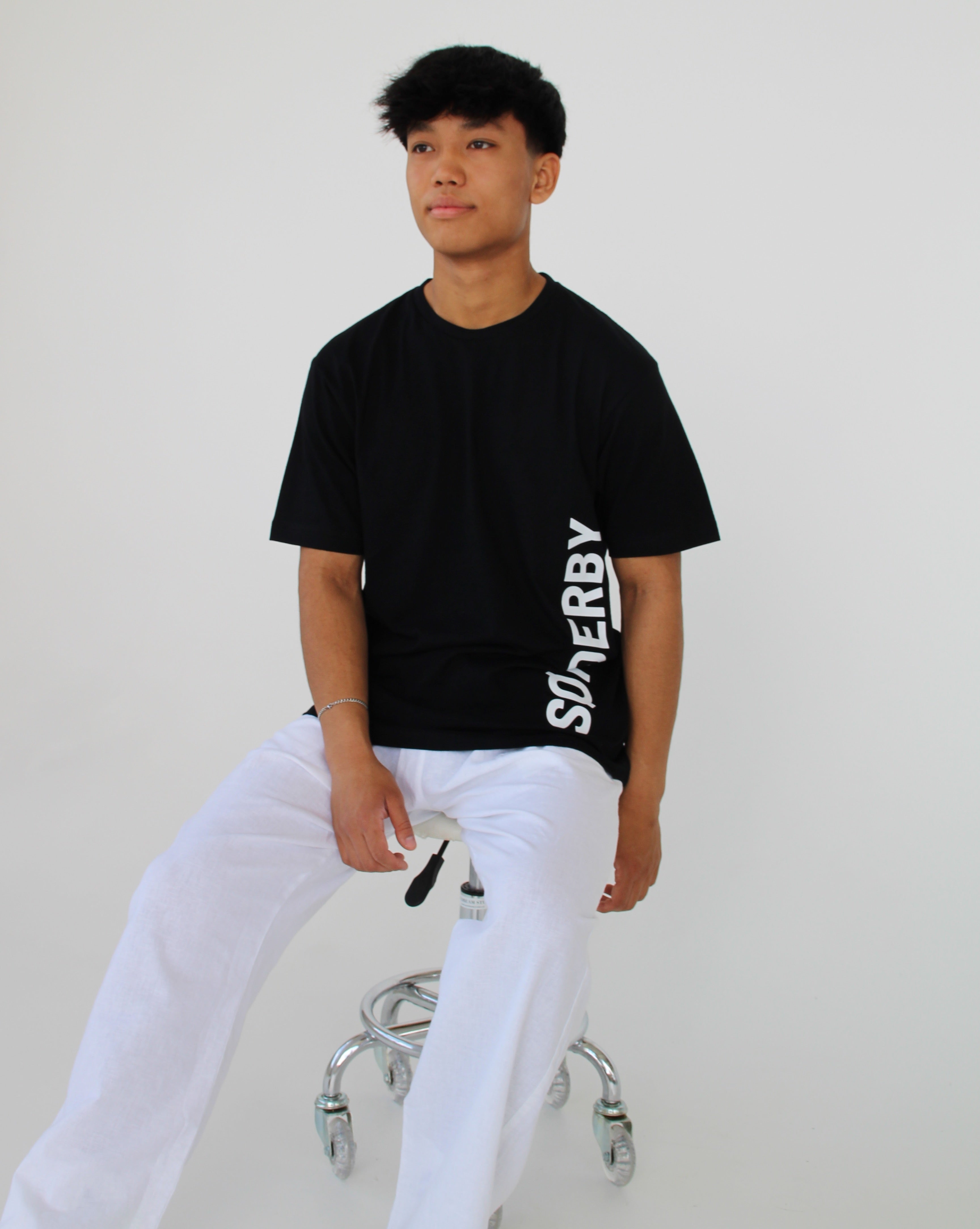En person sidder på en rullende stol, iført en sort T-shirt med ordet "SUNDAY" og hvide bukser, i et hvidt rum.