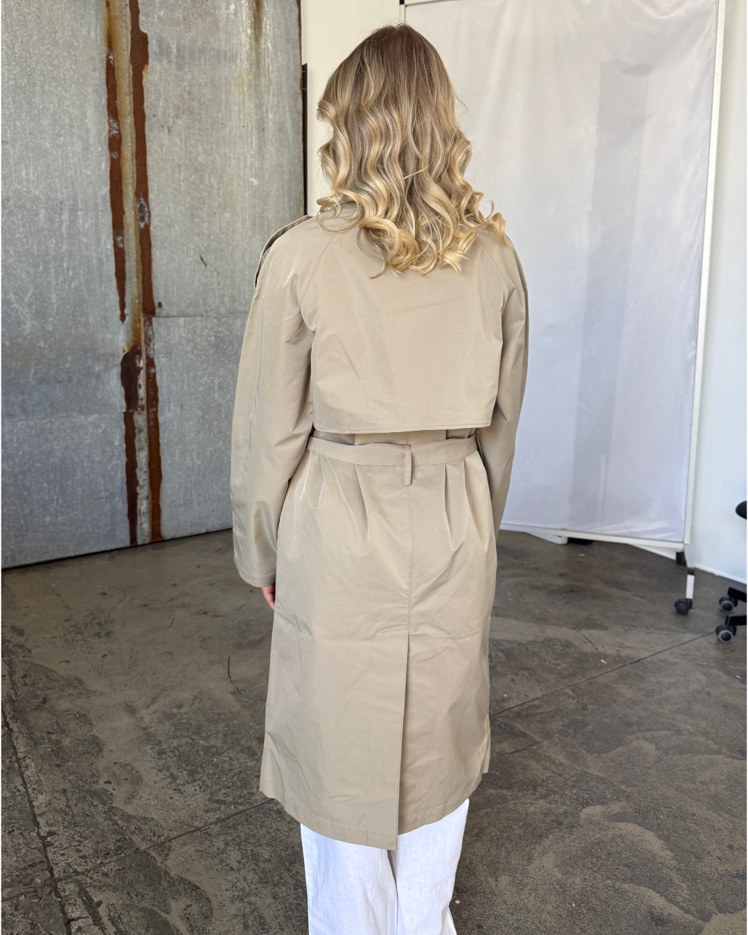 Kvinde med blondt, krøllet hår har en lys beige trenchcoat på. Hun står i et industrielt rum med beton gulv og metalvæg. Trenchcoaten har et bælte i taljen.