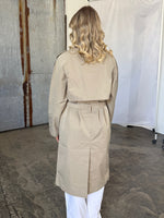 Kvinde med blondt, krøllet hår har en lys beige trenchcoat på. Hun står i et industrielt rum med beton gulv og metalvæg. Trenchcoaten har et bælte i taljen.