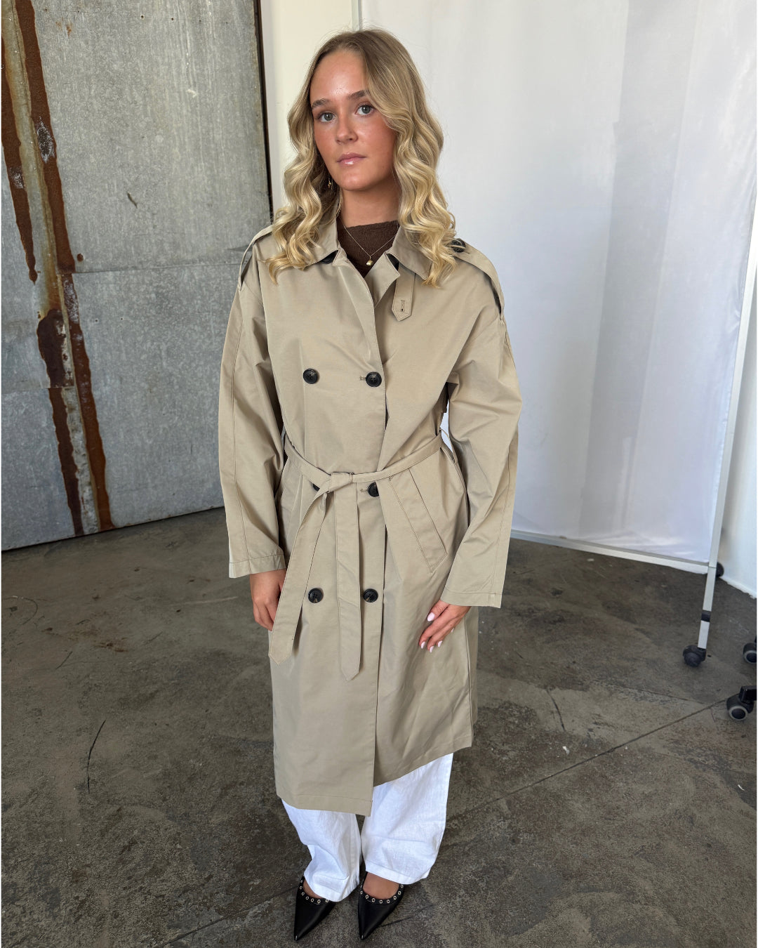 En kvinde står iført en beige trenchcoat med bælte, hvide bukser og sorte sko. Hun er placeret i et fotostudie med beton- og metalbaggrund.