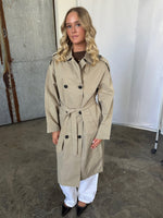 En kvinde står iført en beige trenchcoat med bælte, hvide bukser og sorte sko. Hun er placeret i et fotostudie med beton- og metalbaggrund.