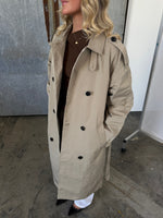 Kvinde iført beige trenchcoat står indendørs på betongulv. Hun har krøllet hår og en brun trøje under frakken. Ved siden af er en metalramme.