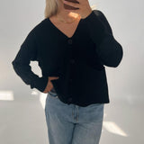 En person iført sort cardigan og lyse jeans tager et spejlfoto. Personen dækker ansigtet med en telefon, og står i et lyst rum.
