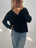 En person iført sort cardigan og lyse jeans tager et spejlfoto. Personen dækker ansigtet med en telefon, og står i et lyst rum.
