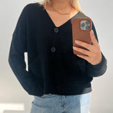 En person iført en sort cardigan med store knapper tager en selfie. De holder en telefon med brunt cover og står i et lyst rum.