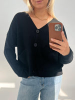 En person iført en sort cardigan med store knapper tager en selfie. De holder en telefon med brunt cover og står i et lyst rum.