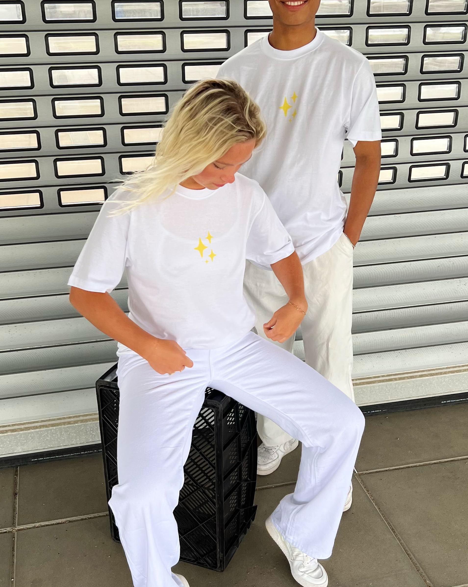 To personer i matchende hvide t-shirts med gule stjerner, står og sidder ved en grå metalskutter. Den ene sidder på en sort kasse, mens den anden står afslappet.