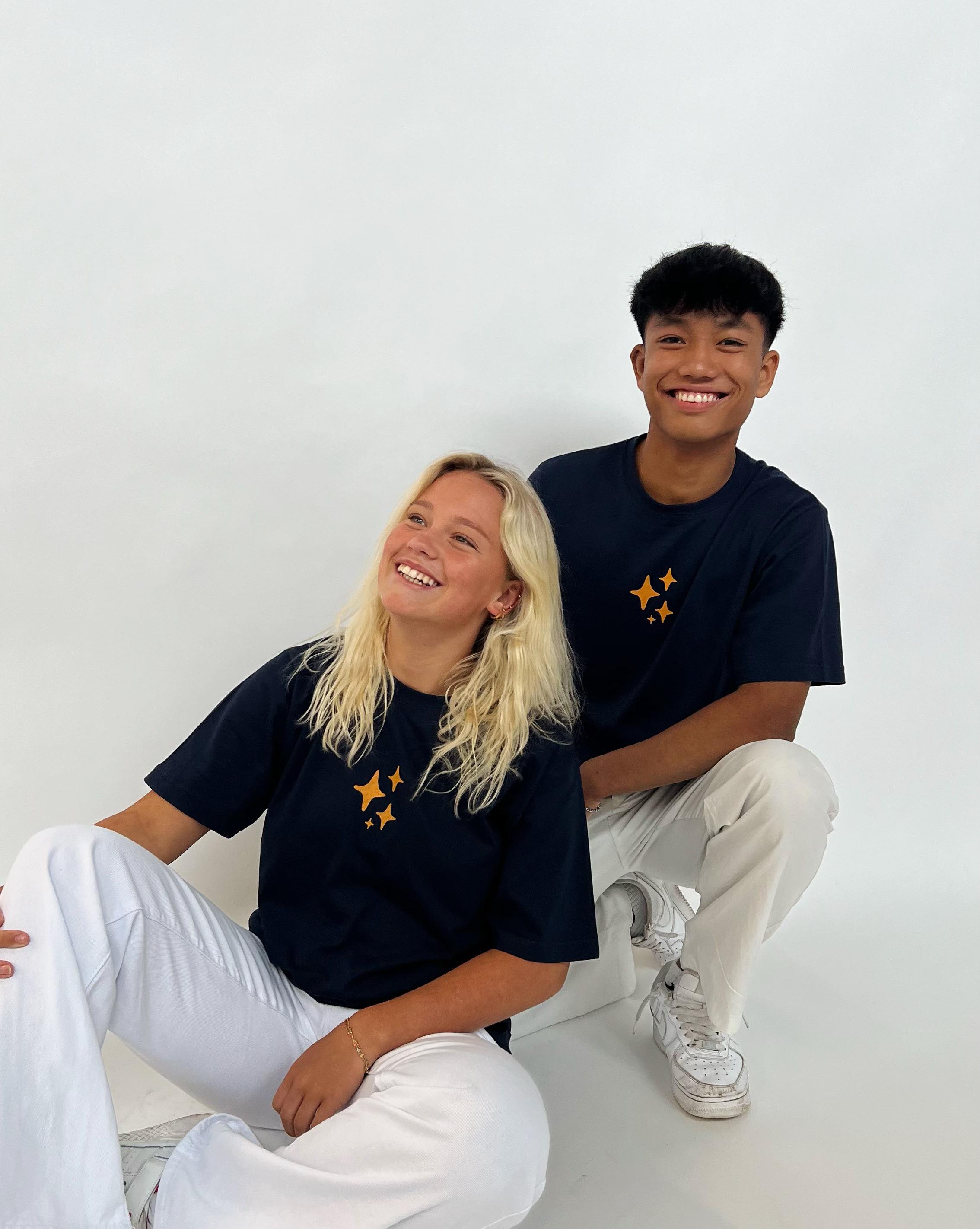 To personer iført mørkeblå t-shirts med orange stjerner sidder og smiler. De sidder på et hvidt gulv i et lyst rum, og begge har hvide bukser på.