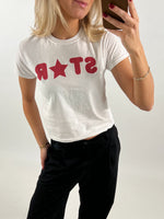 En person iført en hvid T-shirt med rød tekst "STAR" tager et spejlbillede med en smartphone. Personen står i et neutralt, lyst rum.