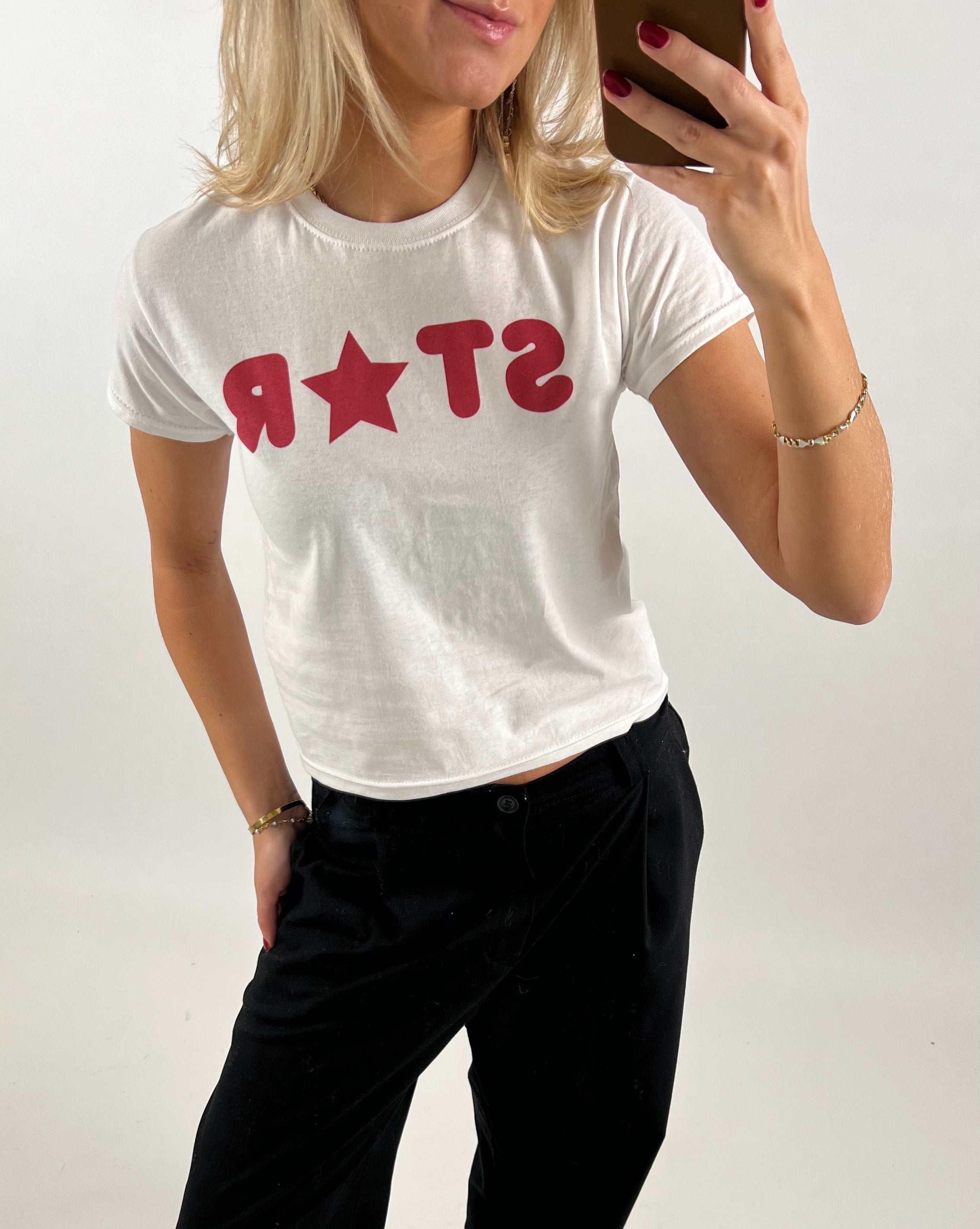 En person iført en hvid T-shirt med rød tekst "STAR" tager et spejlbillede med en smartphone. Personen står i et neutralt, lyst rum.