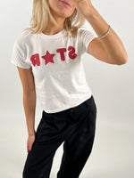 En person iført en hvid T-shirt med rød tekst "STAR," poserer med hånden løftet til ansigtet i en lys indendørs setting. Personen har sort bukser på.