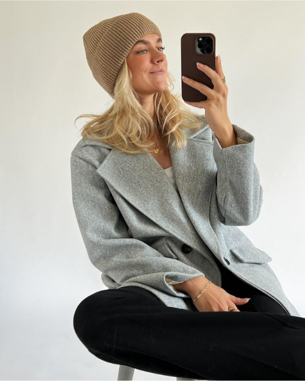 En person sidder og holder en mobiltelefon, iført en grå jakke og beige hue, mod en neutral baggrund.