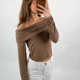 En person tager et spejlbillede iført en off-shoulder, brun top og hvide bukser. Personen holder en brun mobiltelefon, og baggrunden er enkel og hvid.