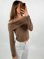 En person tager et spejlbillede iført en off-shoulder, brun top og hvide bukser. Personen holder en brun mobiltelefon, og baggrunden er enkel og hvid.
