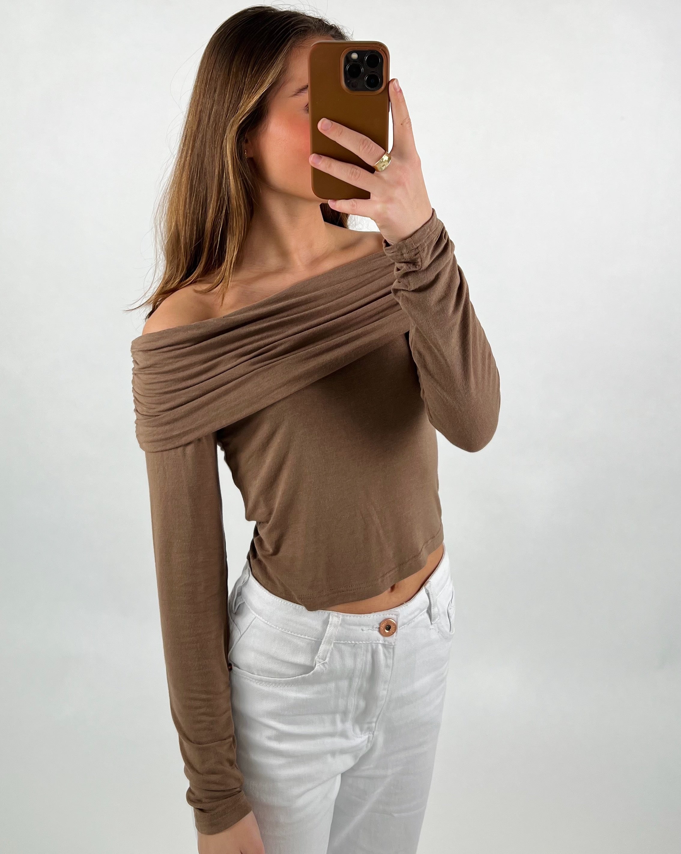 En person tager et spejlbillede iført en off-shoulder, brun top og hvide bukser. Personen holder en brun mobiltelefon, og baggrunden er enkel og hvid.