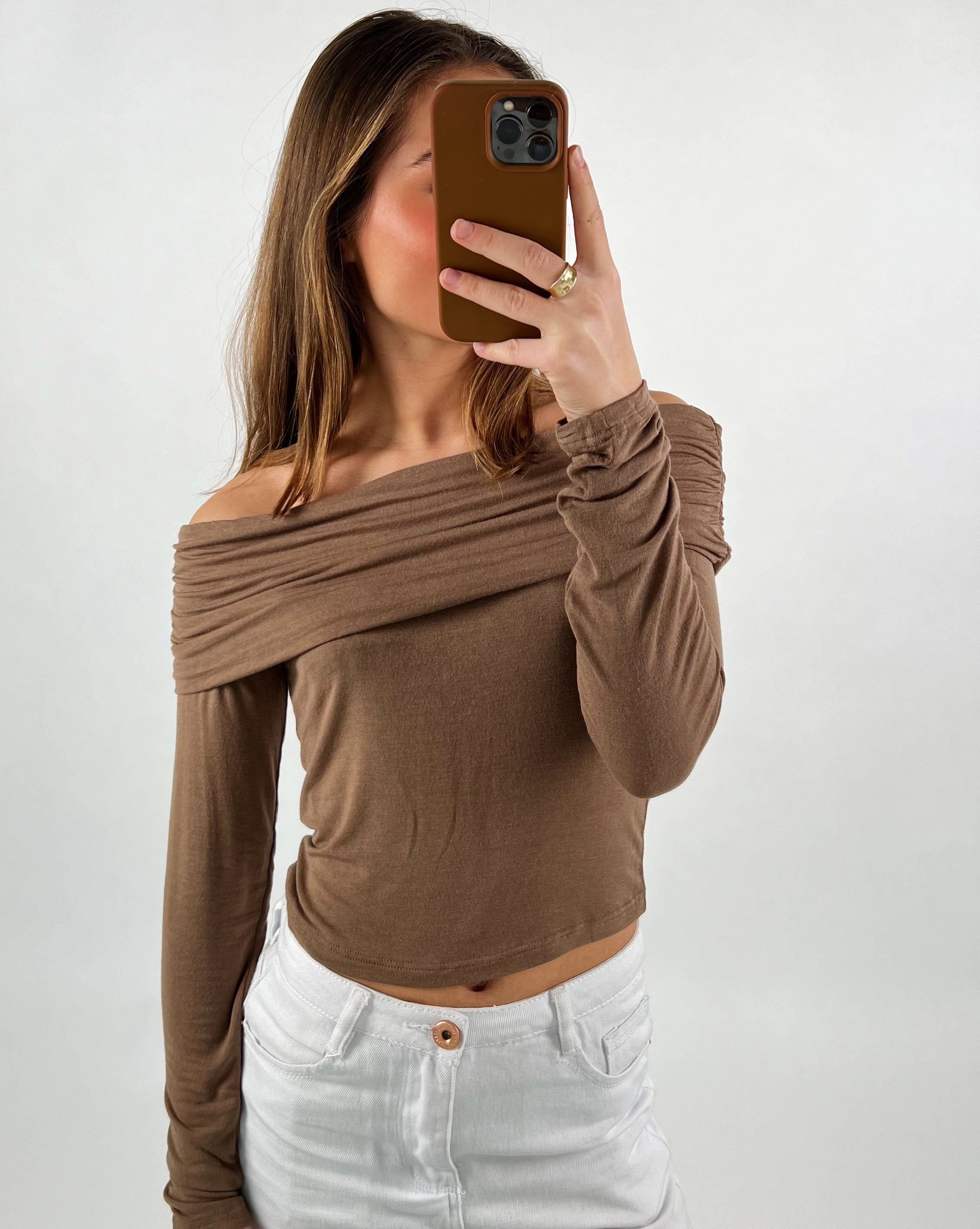Person tager et spejlbillede med en mobiltelefon, iført brun, skulderfri top og hvide bukser. Baggrunden er neutral og lys.
