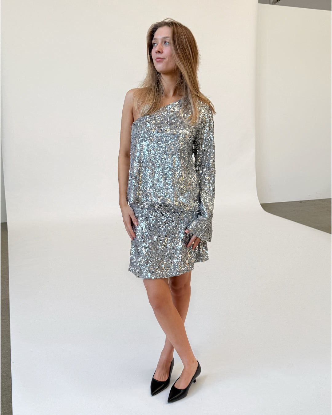 Modelfoto af pige i sølvfarvet TARA one-shoulder bluse og TARA nederdel, med pailetter, samt sorte sko med hæl. Alt er fra BySofieSønderby.