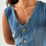 En person iført en denim vest med knapper, bærer to delikate halskæder med små ovale vedhæng. Personen står mod en neutral baggrund.