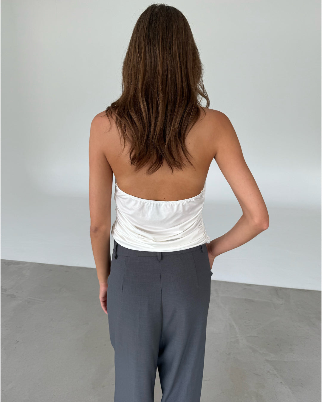 Modelfoto af pige som bærer en hvid TAYLOR halterneck top, samt grå kontorbukser. Set bagfra. Alt er fra BySofieSønderby