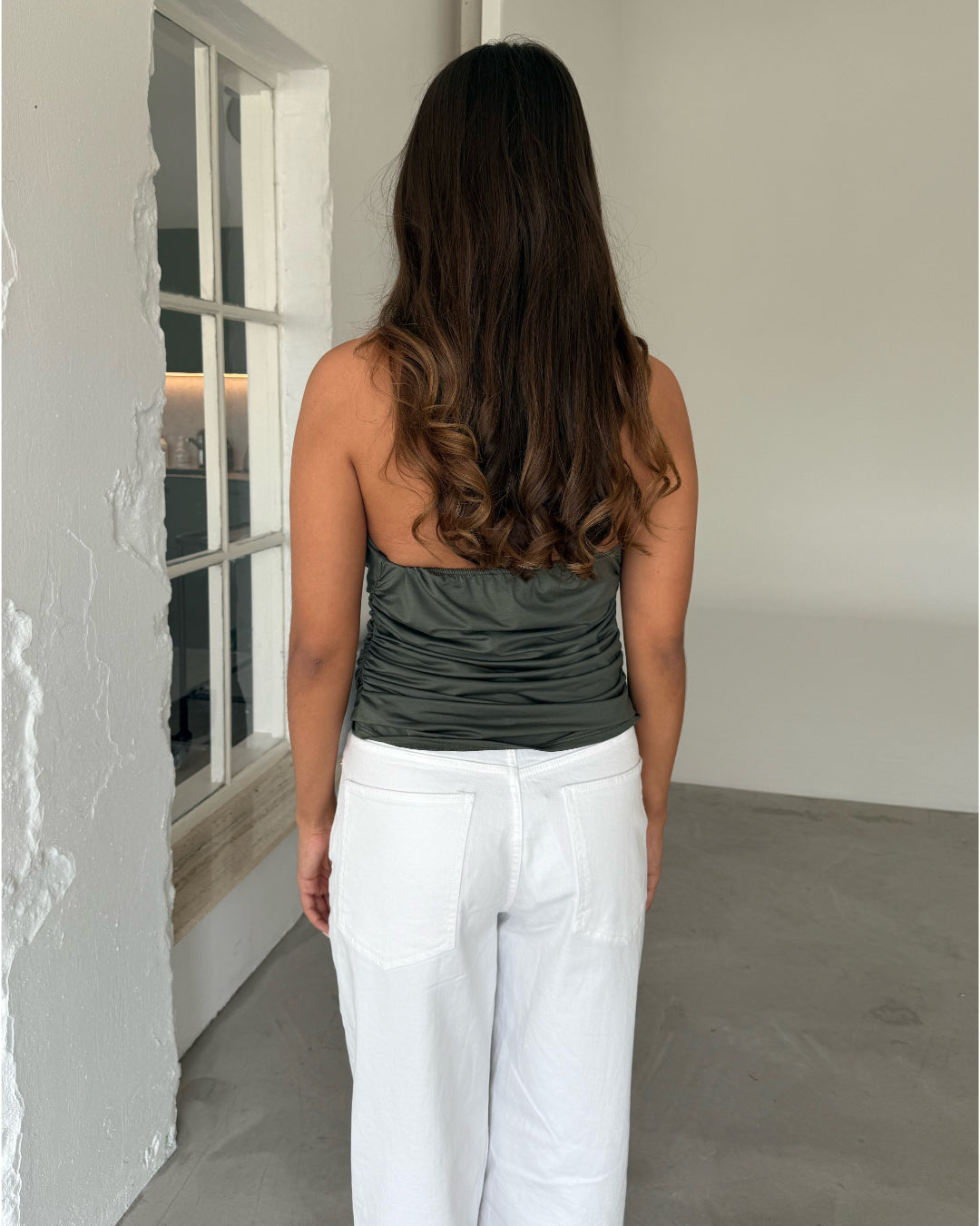 Modelfoto af pige som bærer en kakifarvet TAYLOR halterneck top, samt en hvide jeans. Set bagfra. Alt er fra BySofieSønderby