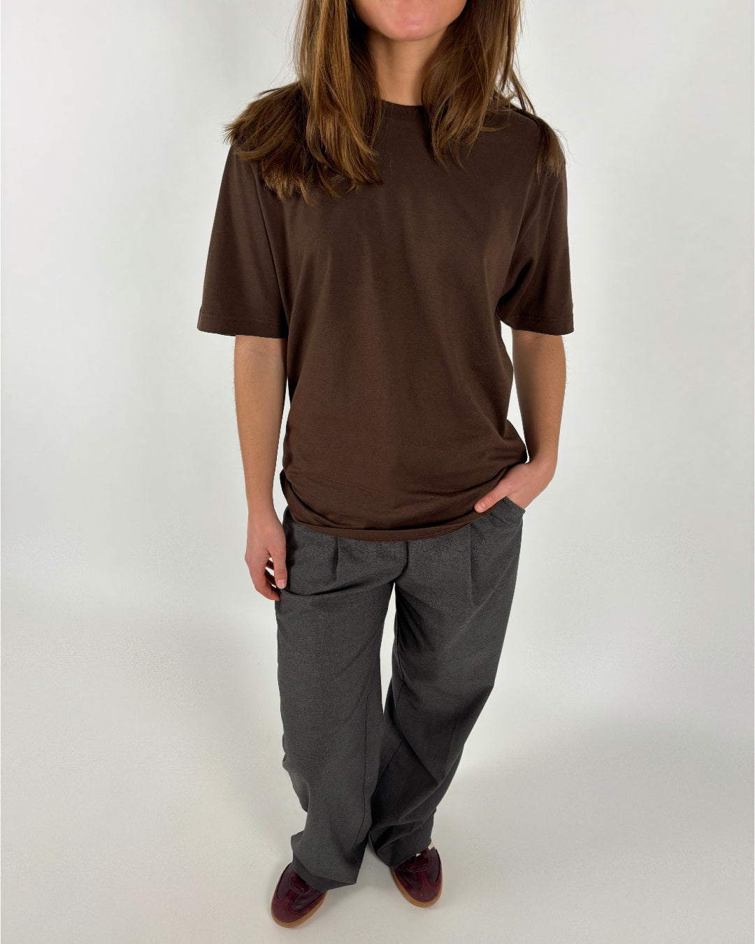 En person står, iført en brun T-shirt og grå bukser, med hånd i lommen, på en lys, neutral baggrund.