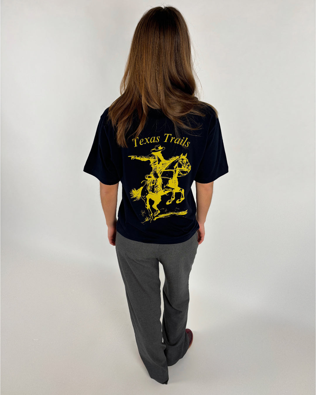 En person med mørkt hår står med ryggen til, iført en sort T-shirt med gul grafik af en cowboy på en hest og teksten "Texas Trails", på en neutral baggrund.
