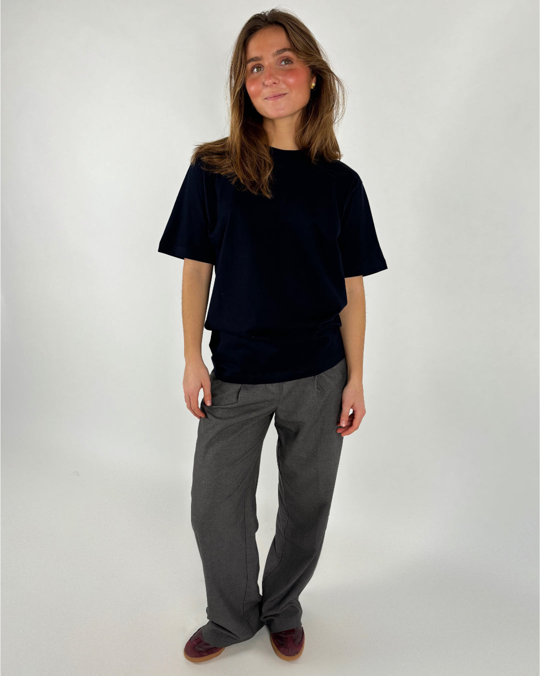 En person står iført en sort t-shirt og grå bukser, smilende let i et neutralt, lyst studieomgivelser. Hun har løst hår og brune sko.