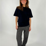 En person står iført en sort t-shirt og grå bukser, smilende let i et neutralt, lyst studieomgivelser. Hun har løst hår og brune sko.