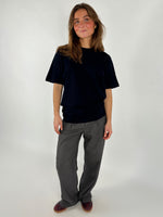 En person står iført en sort t-shirt og grå bukser, smilende let i et neutralt, lyst studieomgivelser. Hun har løst hår og brune sko.