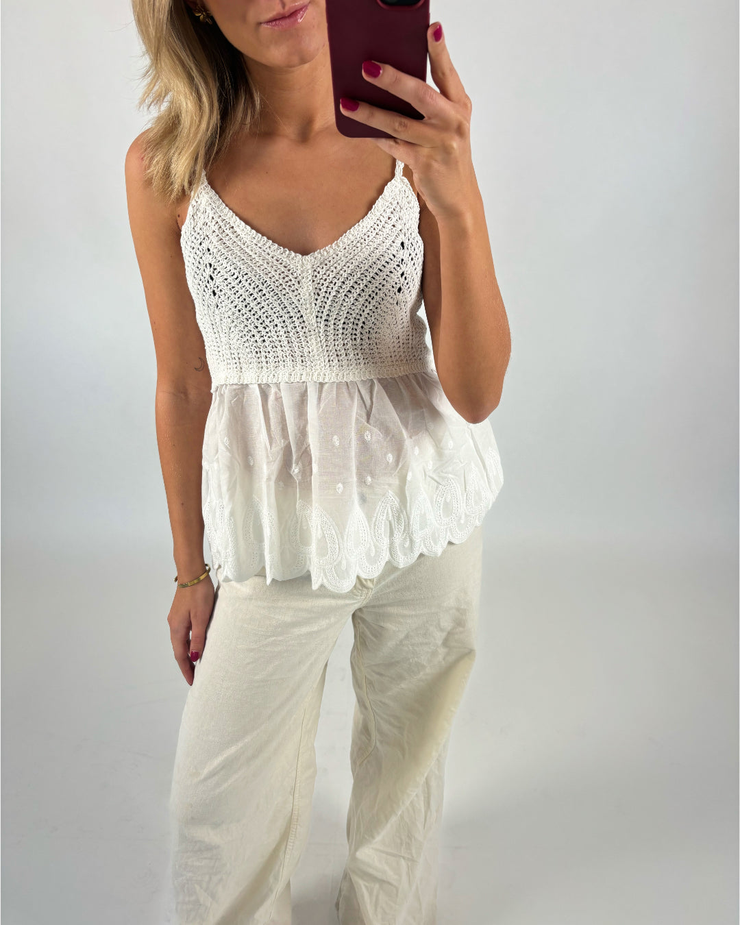 En person iført en hvid, strikket top med blonder, tager en selfie med en rød mobiltelefon i et neutralt lyst rum. Hun har også beige bukser på.